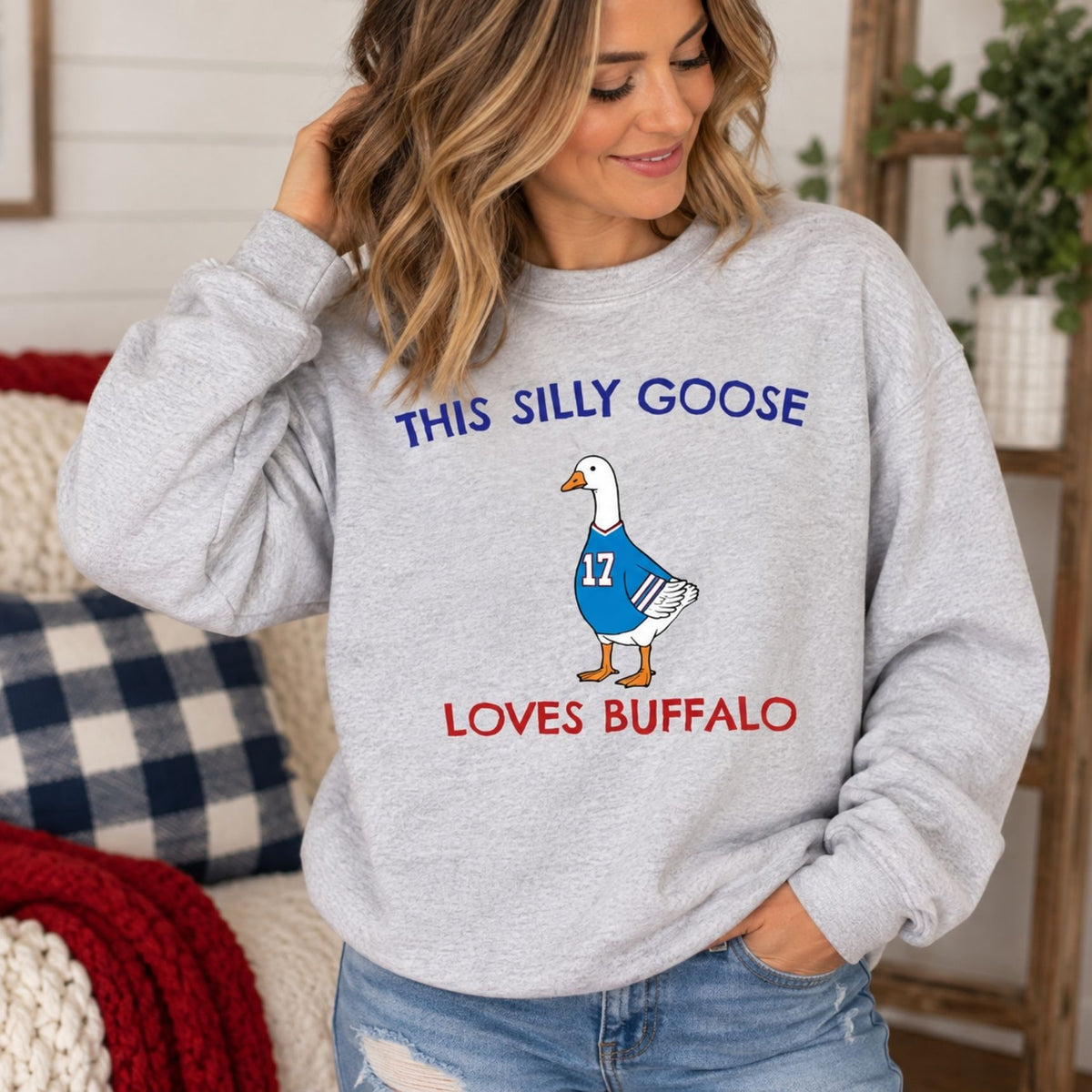 Woman wearing heather gray sweatshirt with "This silly goose loves buffalo" on front and image of goose in a blue jersey with 17 on the front. Standing in front of a sofa with blankets.