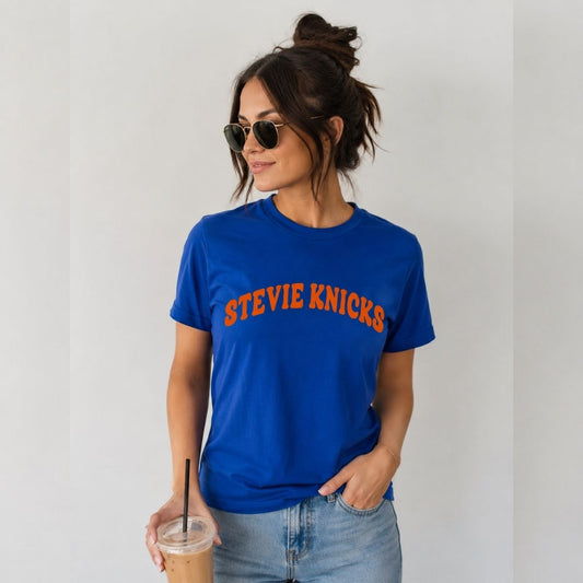 Woman wearing royal blue and orange "Stevie Knicks" tshirt inspired by Taylor at NY Basketball game, holding a latte on a gray background.