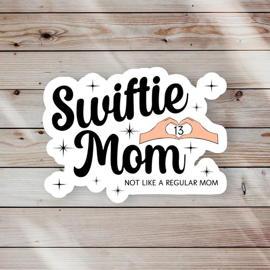 white sticker with black text "Swiftie Mom" "Not like a regular mom" 13 heart hands -waterproof sitting on wood background. Little Gift Nook