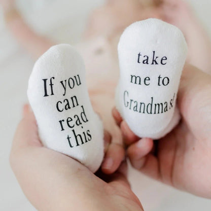 white baby socks on baby's feet with black text on bottoms "If you can read this" "take me to Grandma's" being held up by woman's hands.
