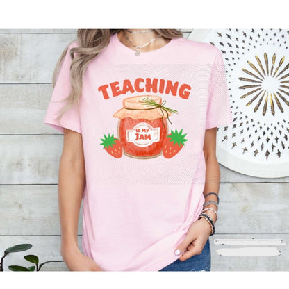 Woman wearing pink tshirt with orange/red jam jar, strawberries, and "Teaching is my jam" on front on a wood plank background. Little Gift Nook