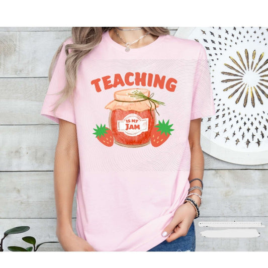 Woman wearing pink tshirt with orange/red jam jar, strawberries, and "Teaching is my jam" on front on a wood plank background. Little Gift Nook