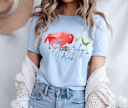 woman wearing light blue tshirt with red buffalo, wine glasses, and text "This is why I drink" image on front. Standing on gray background with cardigan off shoulders. 