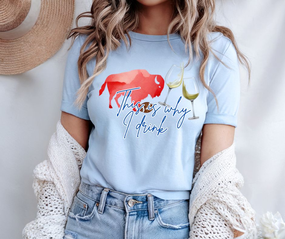 woman wearing light blue tshirt with red buffalo, wine glasses, and text "This is why I drink" image on front. Standing on gray background with cardigan off shoulders. 