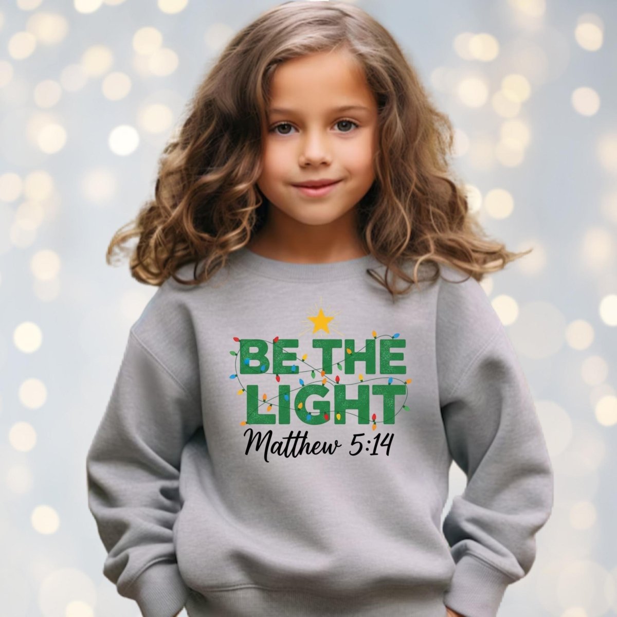 Child wearing gray BE THE LIGHT Christmas lights Matthew 5:14 sweatshirt on blurry lights background.