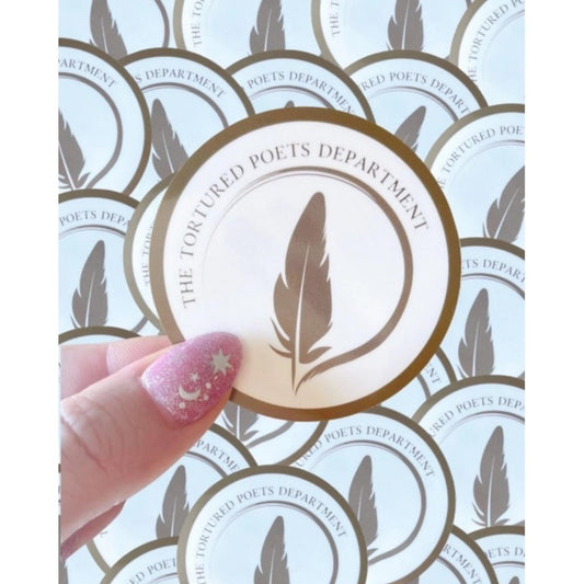 hand holding round beige and chrome colored sticker with feather and "The Tortured Poets Department" on front on background of more stickers.