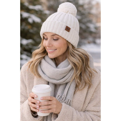 Woman wearing a white knit beanie with a pom-pom hat and a gray scarf, holding a white cup outdoors in winter.
