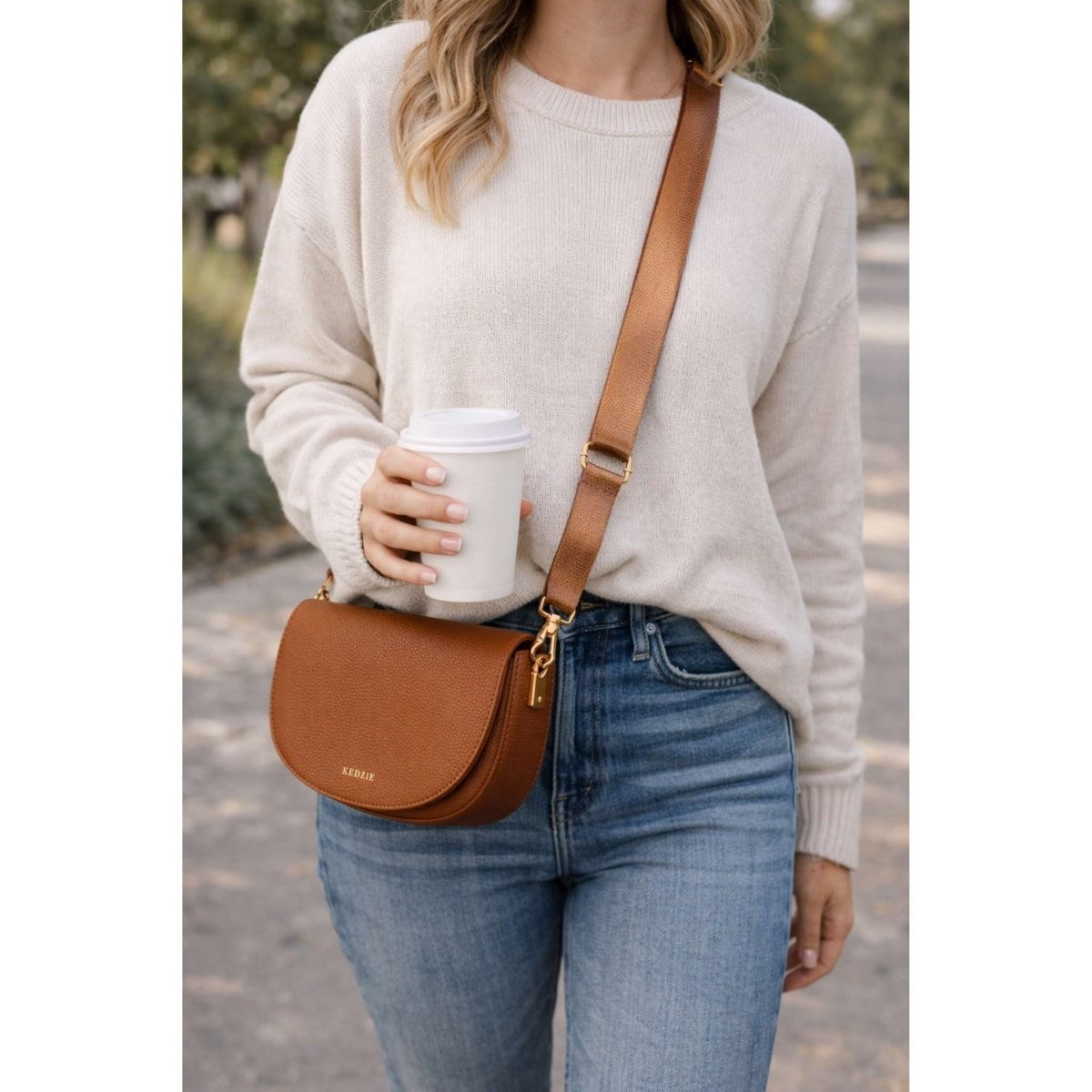 Woman wearing a beige sweater, blue jeans, and carrying a chestnut brown Kedzie lune crossbody bag purse outdoors.