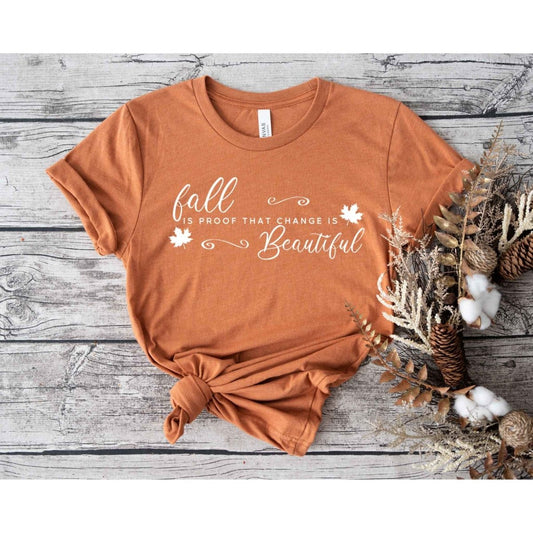 Autumn orange "fall is proof that change is beautiful" white lettering tshirt on wood  background with leaves. Little Gift Nook
