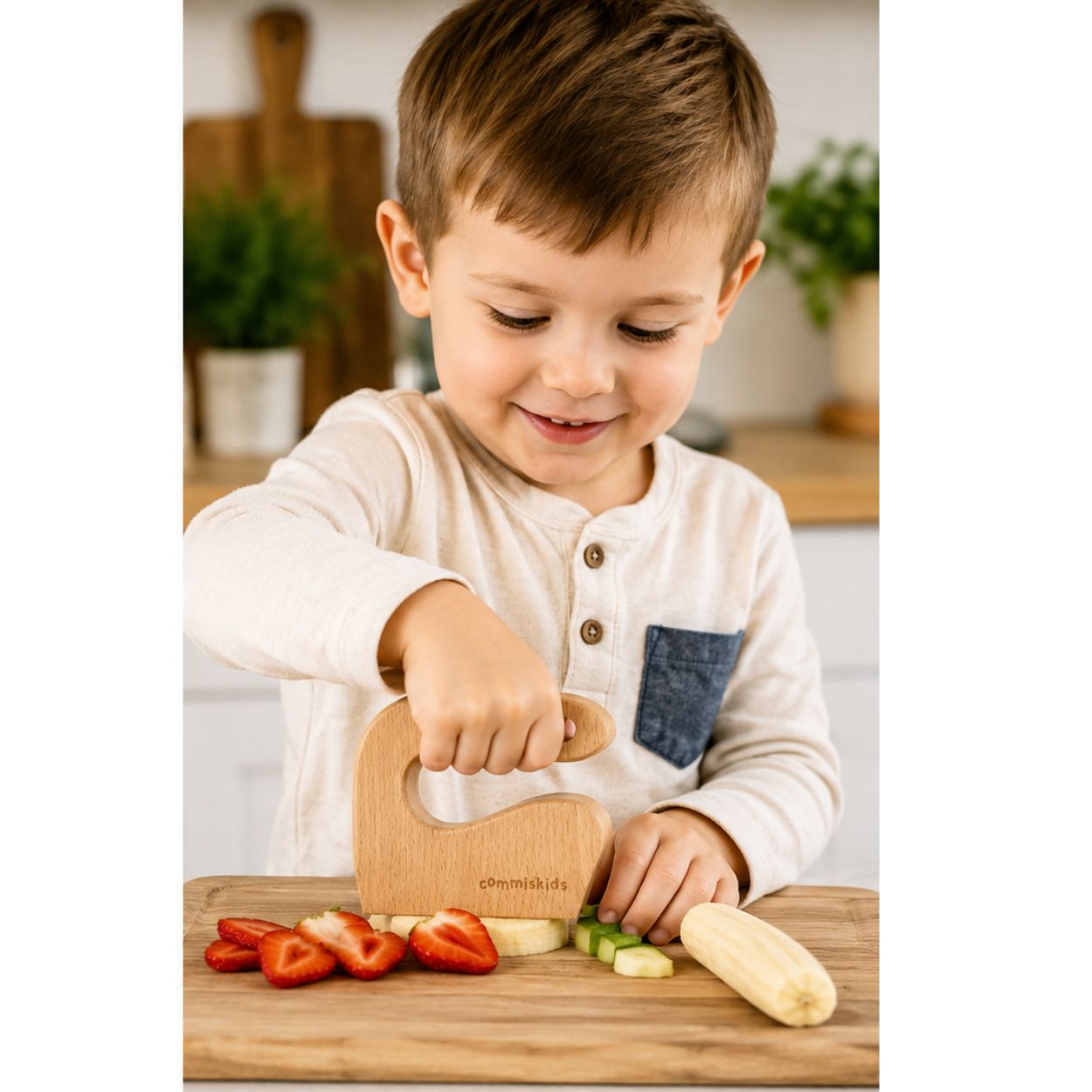 A young boy with short brown hair is smiling and cutting vegetables on a wooden board using a wooden chopper. He is wearing a cream-colored long-sleeve henley shirt with a blue pocket.