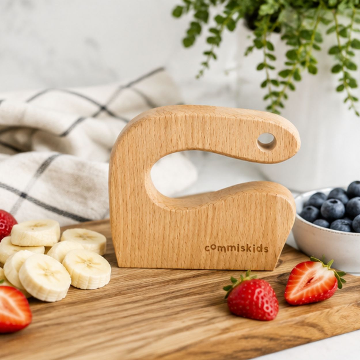 A wooden letter C kids chopper displayed on a wooden cutting board surrounded by sliced bananas, strawberries, and blueberries.