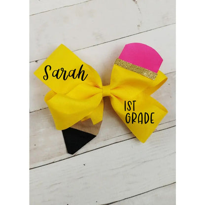 A personalized yellow and pink pencil them hair bow with black text reading 'Sarah' and '1st Grade', displayed on a white wood plank background.
