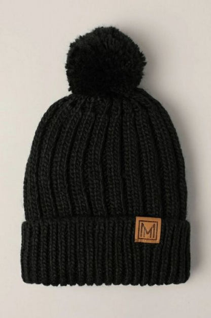 Black knit beanie with pom-pom and brown leather patch laying on a light gray background.