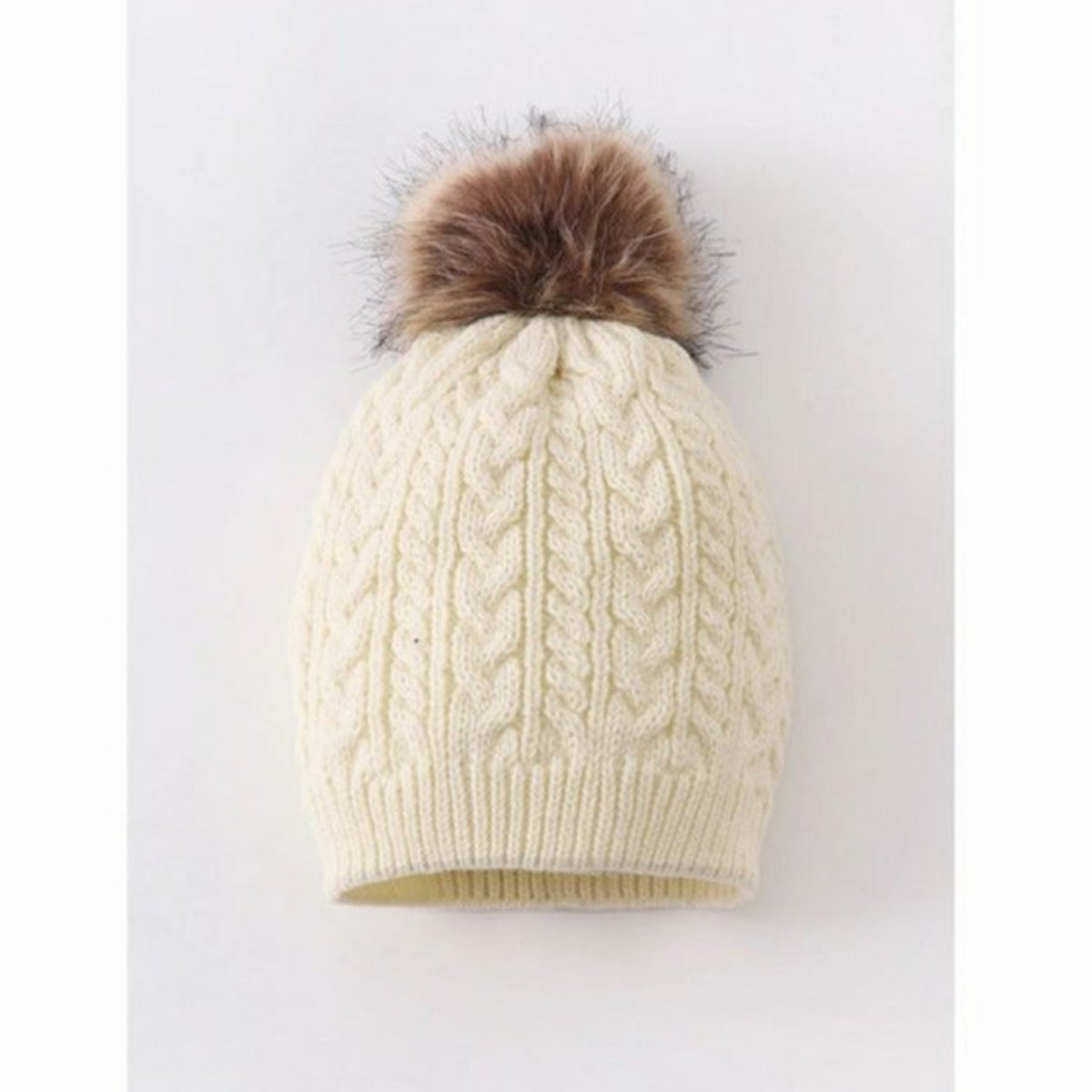Kids creme ivory cable knit bean hat with tan faux fur pom at top sitting on gray and white background. Little Gift Nook