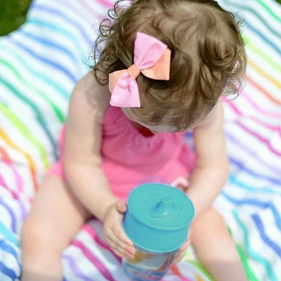 Little Girl using GoSili silicone teal sippy cup lid on a glass on a rainbow blanket background. 