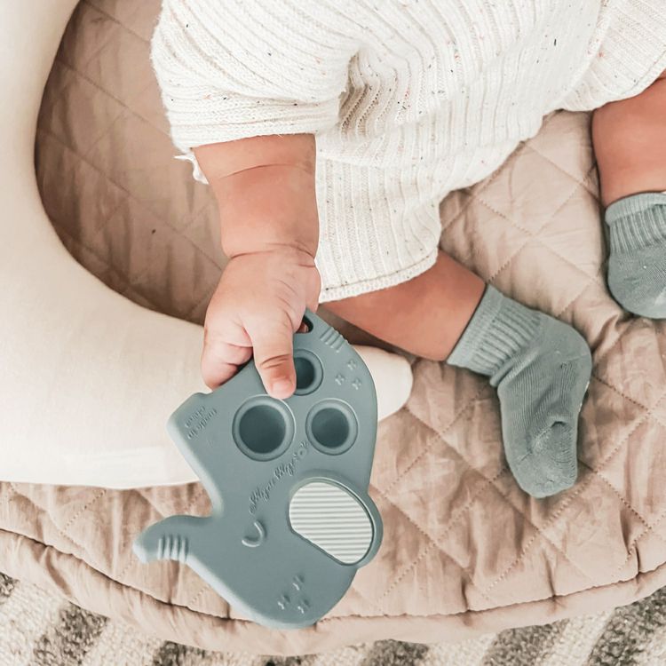 Baby's hand holding Itzy Pop blue elephant teether and popper toy on a beige blanket.