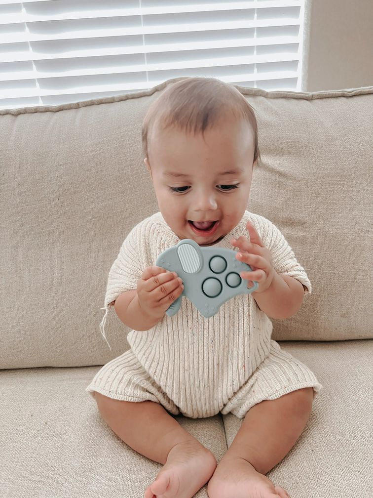 Baby holding blue elephant Itzy Ritzy Itzy Pop teether and popper toy on a beige couch.