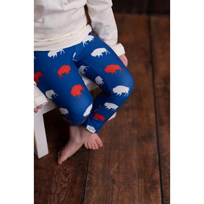 close up of little girl sitting wearing royal blue, red, and white buffalo print leggings on wooden background.
