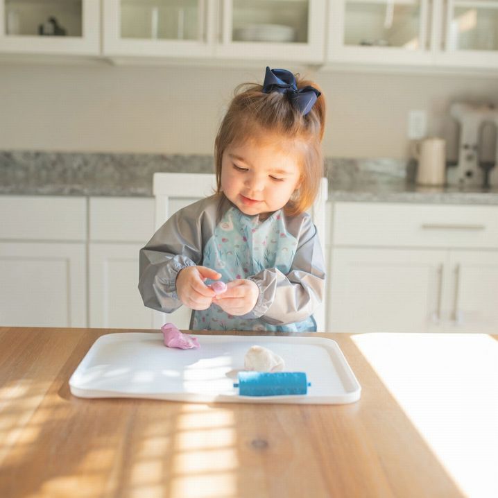toddler girl sitting at table playing with doh wearing a long sleeved wipe off bib. Little Gift Nook