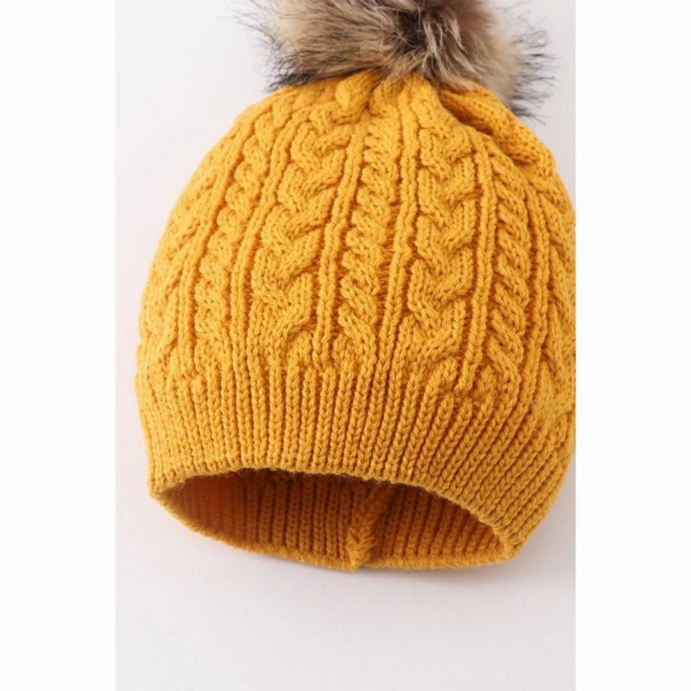 close up of cable knit on kids mustard pom beanie hat sitting on gray and white background.