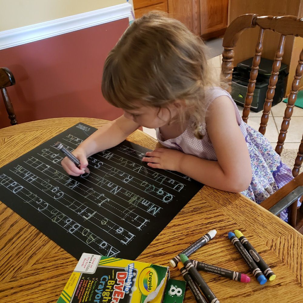 Child using crayons and black reusable plastic letters practice place mat on wooden table.