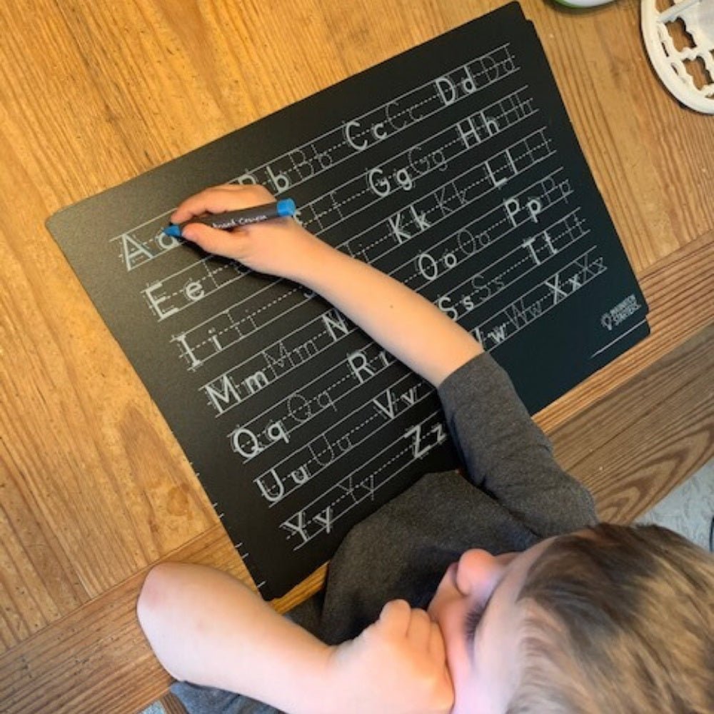 child tracing letters on black wipe off reusable letters practice plastic place mat with crayons on wooden table.