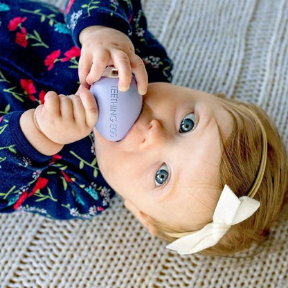 Baby using The Teething Egg lavender egg shaped baby teether on a beige blanket background. Little Gift Nook