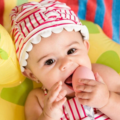 Baby using The Teething Egg pink egg shaped baby teether on a bright background. 